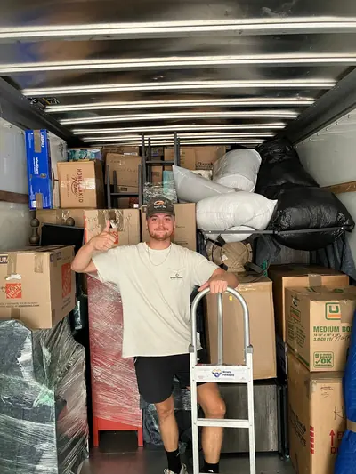 A mover with a hand truck in front of boxes and wrapped furniture