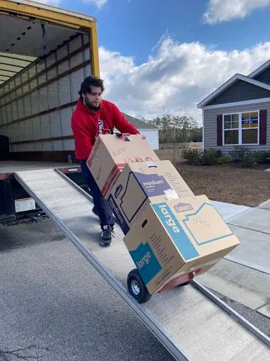 A mover wheeling stacked boxes down a truck ramp on a hand truck