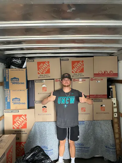 A mover in a UNCW shirt giving two thumbs up in front of boxes stacked to the ceiling of a truck